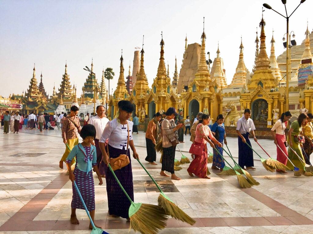 Amidst the hustle and bustle of a crowd, volunteers meticulously sweep the vast marble terrace with large round brooms against a backdrop golden pagodas