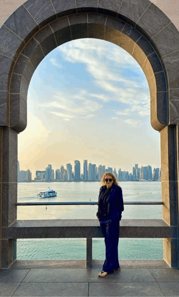 A woman standing on a walkway with blonde hair and sunglasses and city skyline across the water